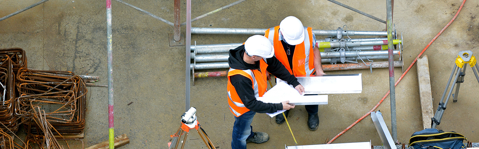 Two construction workers reviewing plans, steal pipes nearby, aerial view